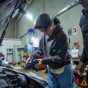 A young person holding a tool and watching attentively as a mechanic in overalls inspects a brake disc on a car raised on a hoist